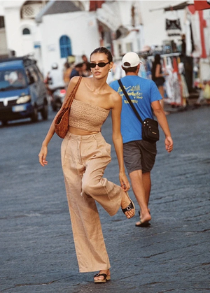 A woman strides confidently on a paved street in a coordinated tan crop top and slacks, with a man walking in the opposite direction.