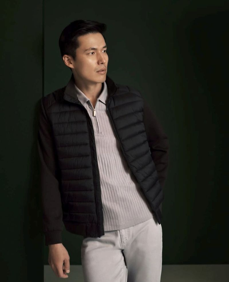 A stylish Asian man poses in a puffer vest over a polo shirt and casual pants against a dark green background in a studio shot.