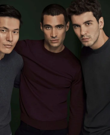 Three diverse young adult men pose indoors in a close-up shot, wearing sweaters against a blurred dark green background, looking at camera.