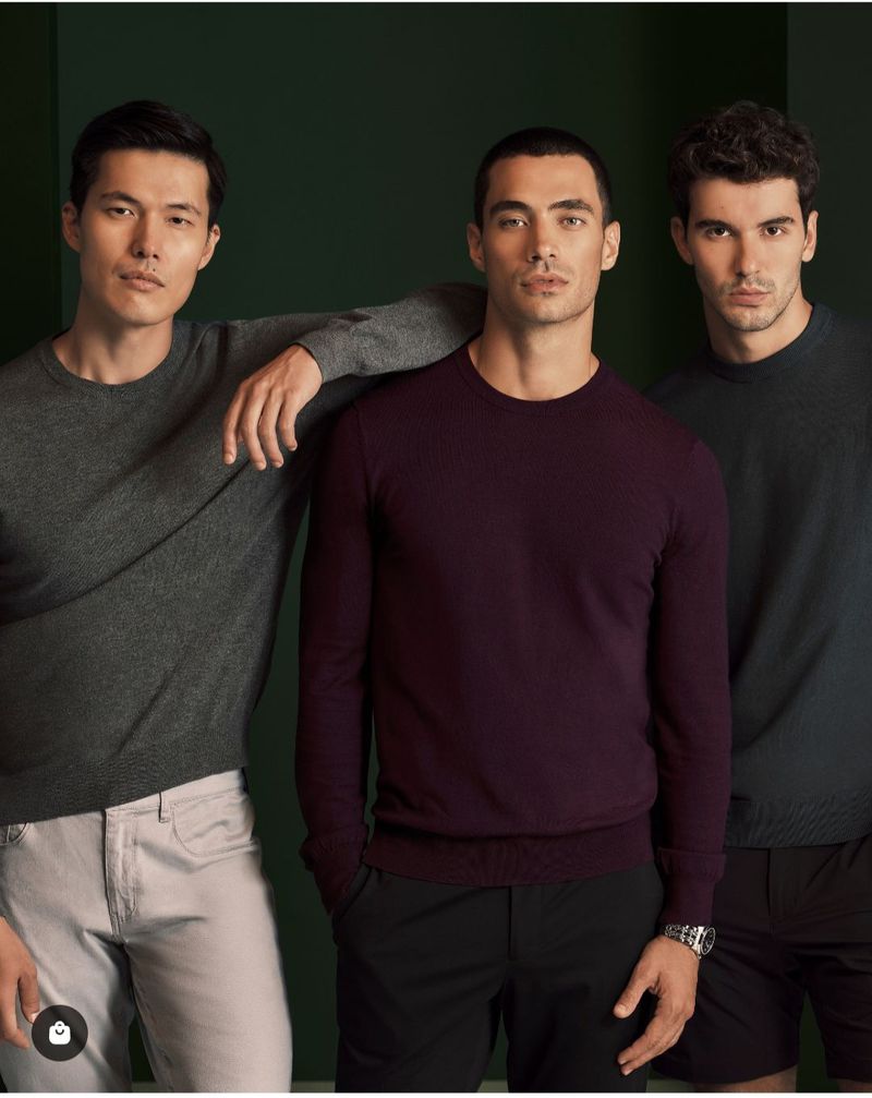 Three adult male fashion models pose indoors against a solid dark-green background. The men are wearing casual pullovers and trousers.
