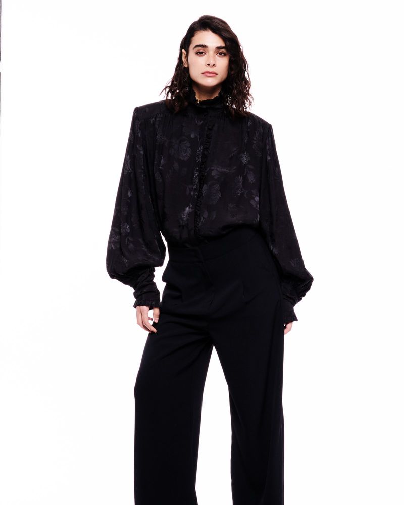 A woman in a black, long-sleeved floral blouse and wide-leg trousers stands in a brightly lit studio, showcasing a fashion-forward ensemble.
