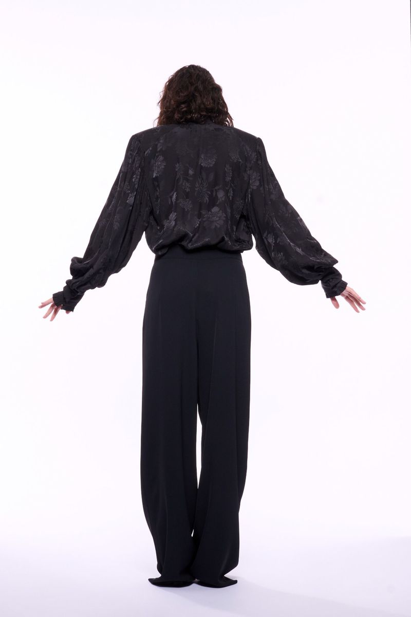 Back view of woman in black patterned long-sleeve blouse with wide-legged pants, standing with arms outstretched against a white backdrop.