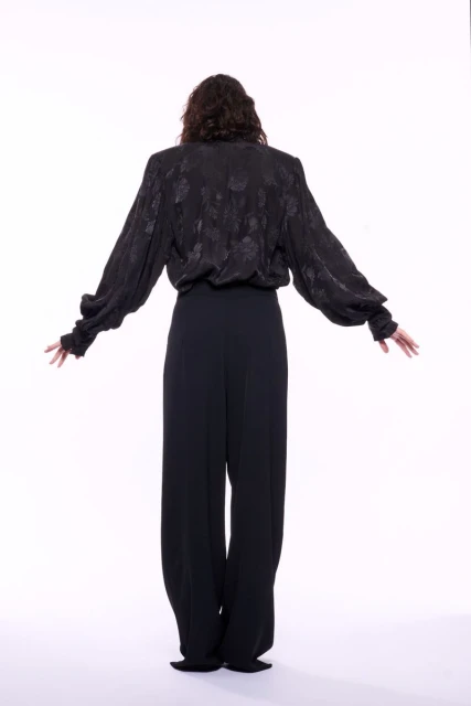 Back view of woman in black patterned long-sleeve blouse with wide-legged pants, standing with arms outstretched against a white backdrop.