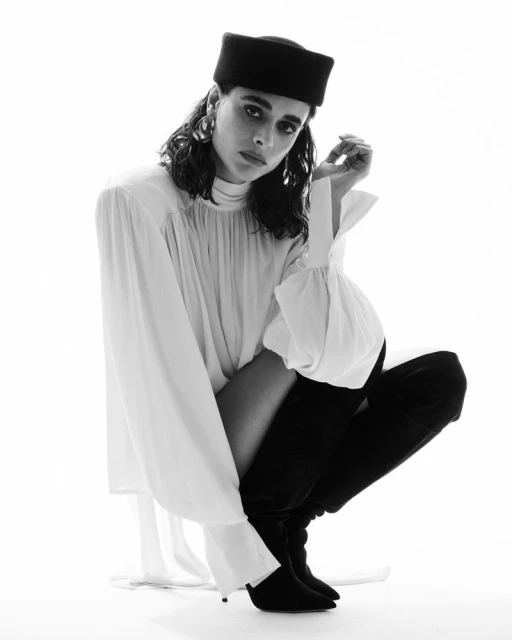 Black and white portrait of a woman squatting in a white dress, black hat, and black boots in a bright, minimalist studio setting.
