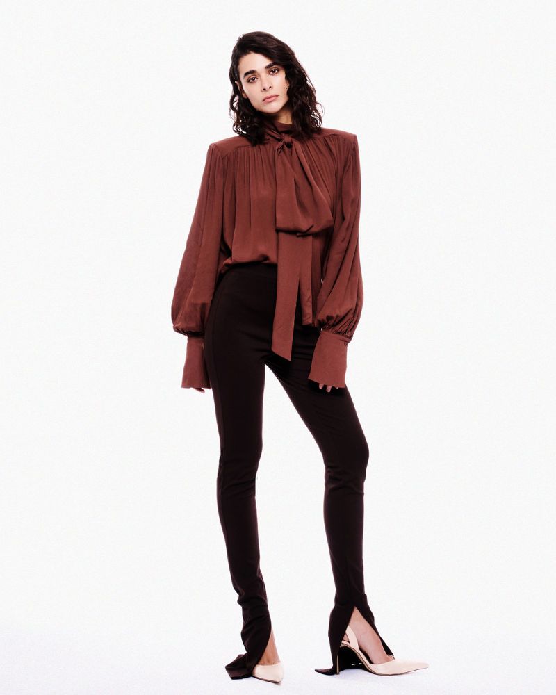 Fashion shot of a woman with long brown hair wearing a brown satin blouse and black pants against a white background in studio lighting.