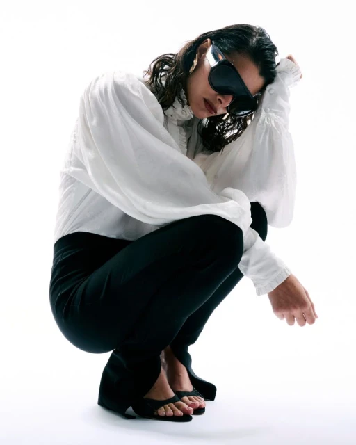 A woman in sunglasses and a white blouse poses while crouching against a white backdrop, creating a high-key, fashion-forward portrait.