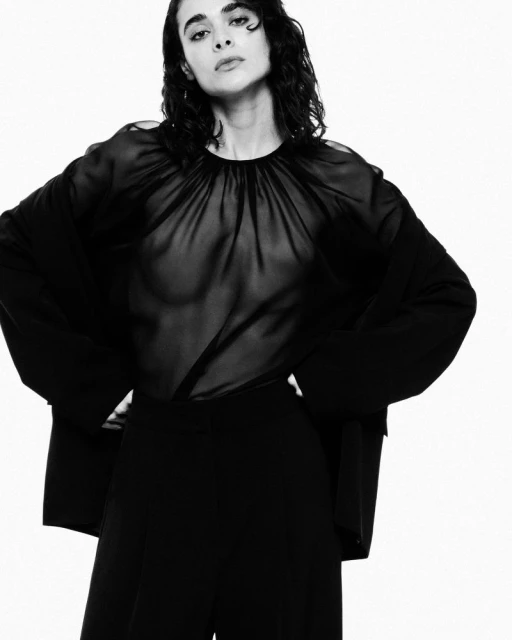 A woman poses in a studio wearing a sheer black blouse, black pants, and a black jacket against a white background in a fashion-style photograph.