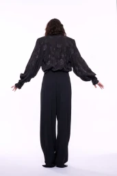 Rear view of a model in a black jacquard blouse with puffed sleeves and wide-leg black trousers against a white seamless background.