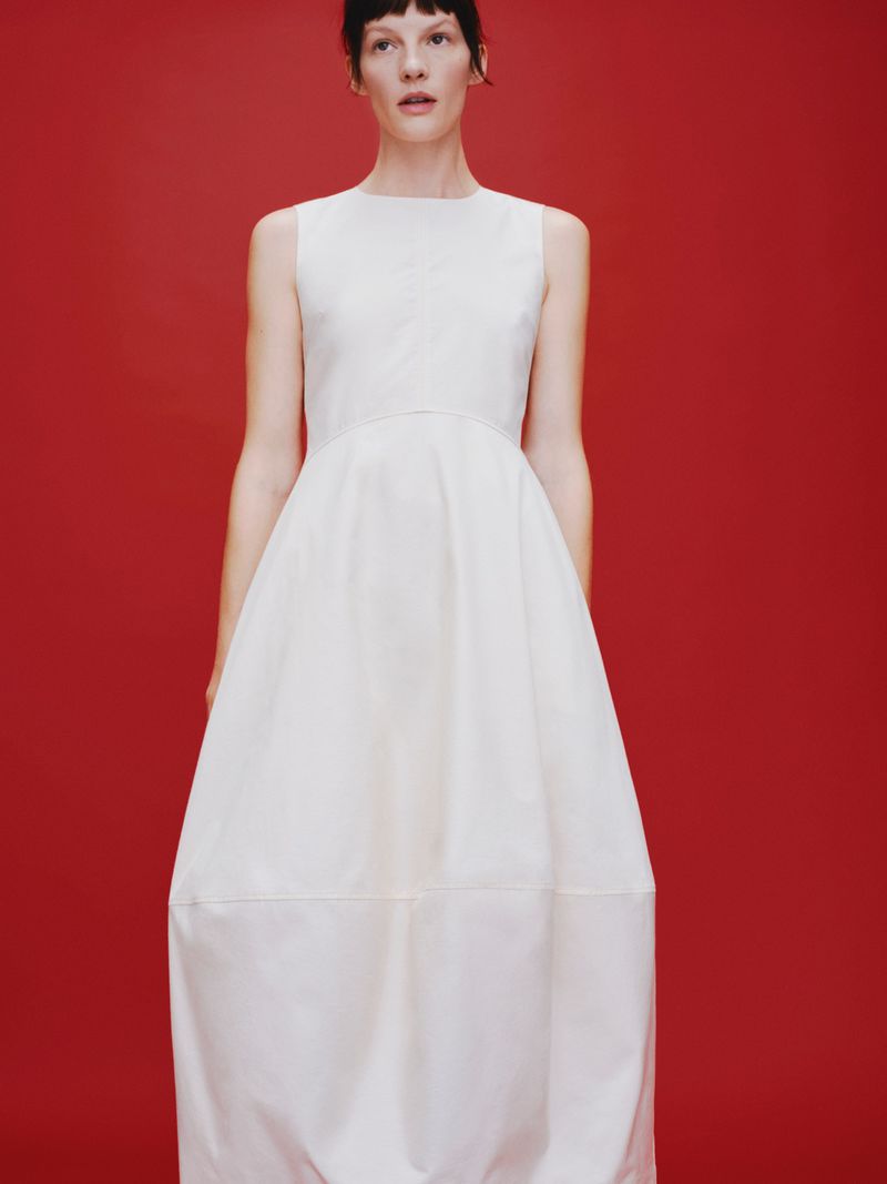 A stylish adult female poses in a full-length, sleeveless white dress against a solid red studio backdrop, showcasing modern fashion.