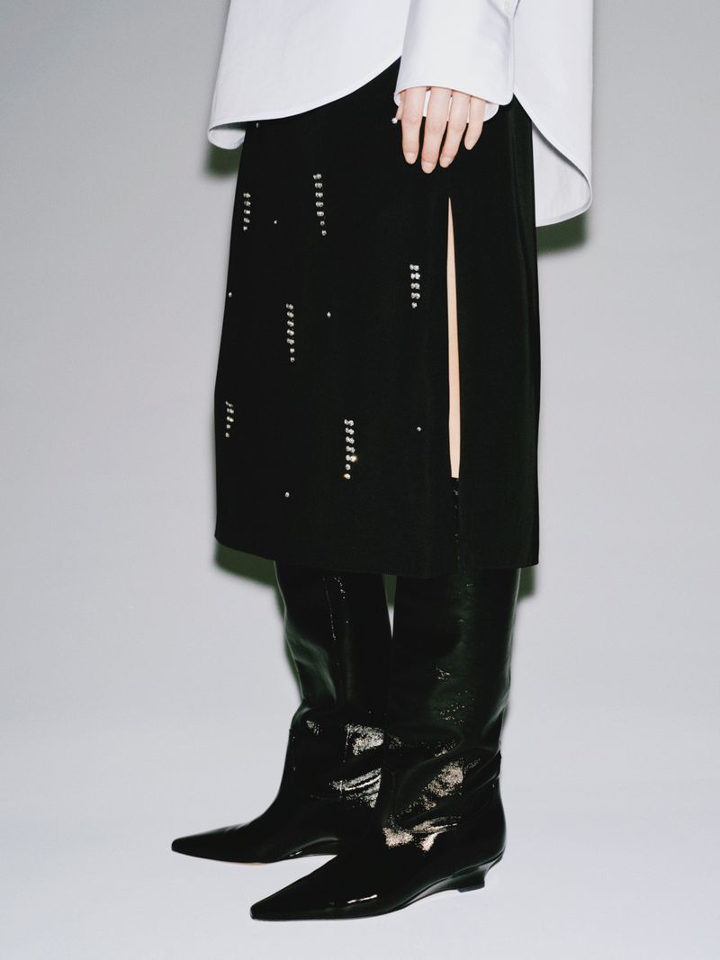 Fashion shot of a person wearing a long-sleeved white shirt, a black skirt with embellishments, and shiny black knee-high boots.