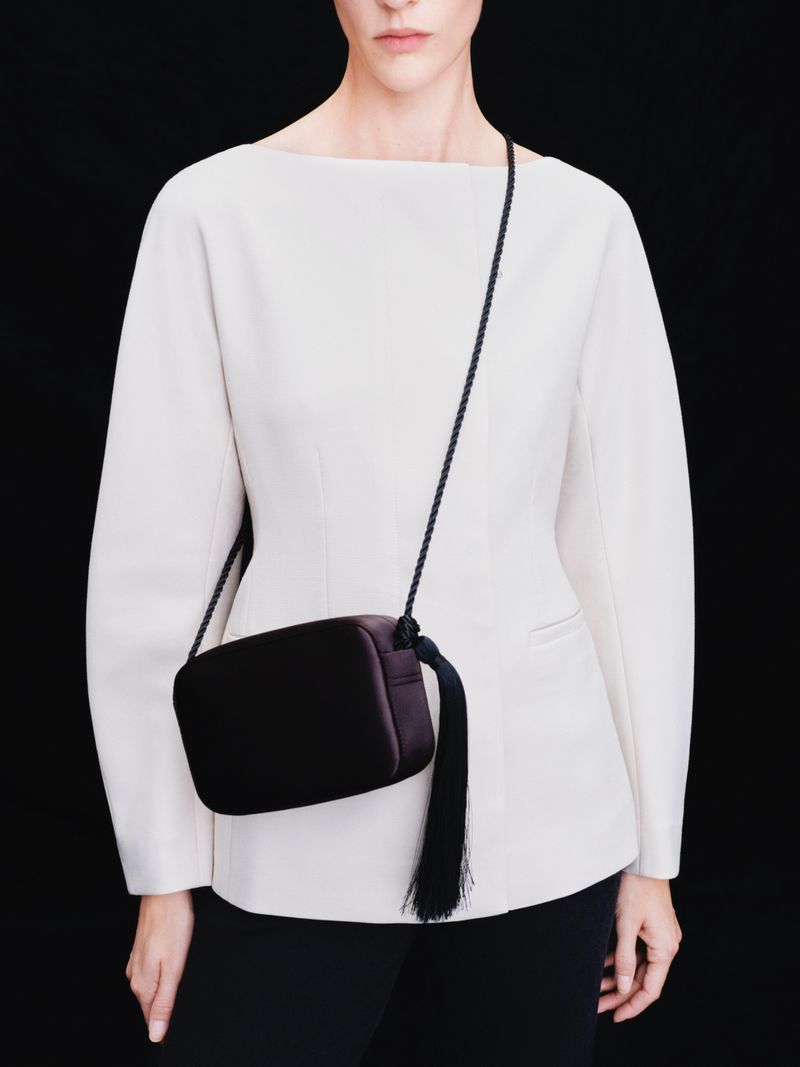 Woman wearing a white jacket and black skirt, showcasing a black crossbody bag with a tassel strap, against a solid black background.