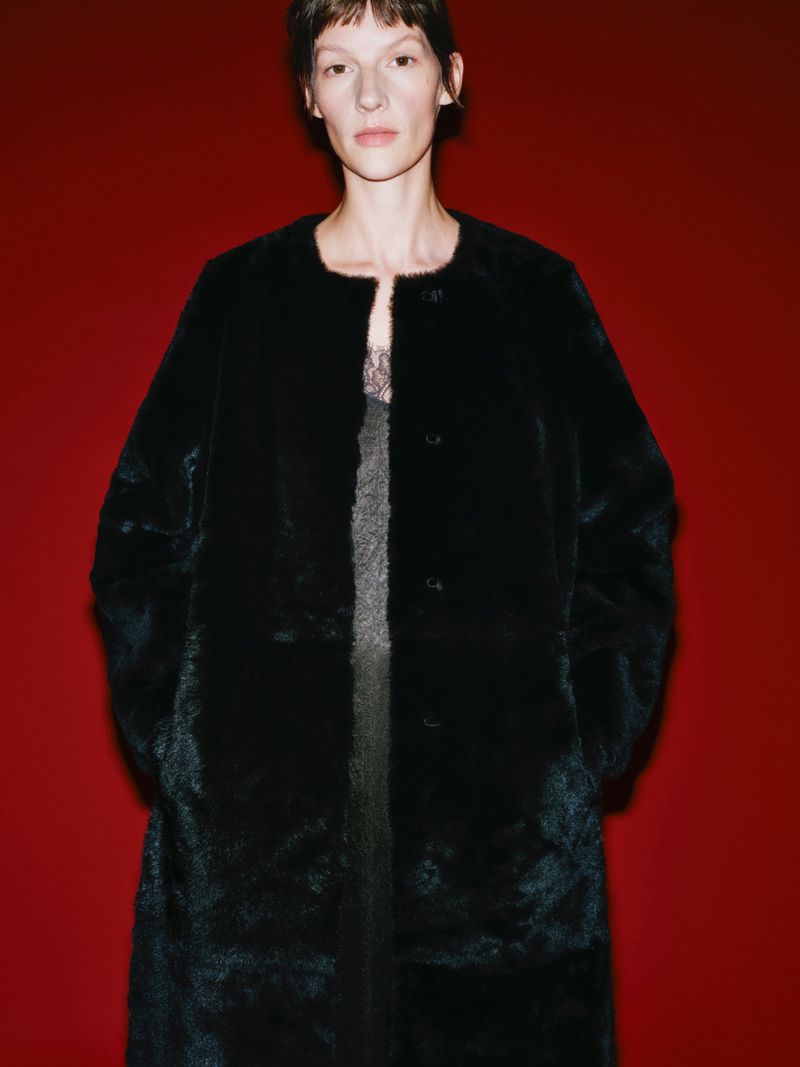 A woman with short brown hair poses in a dark faux-fur coat against a red backdrop in a fashion photography studio setting.