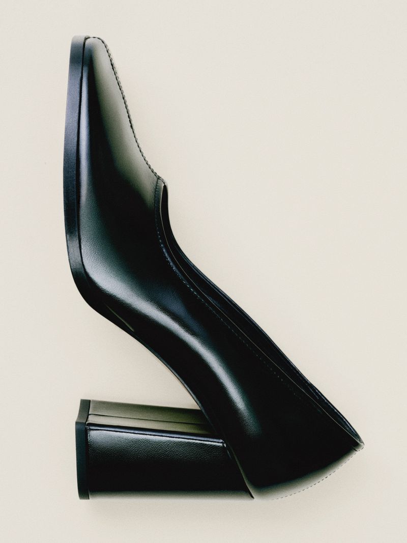 Black leather heeled pump with a block heel on an off-white background. A stylish and modern shoe perfect for fashion-forward individuals.