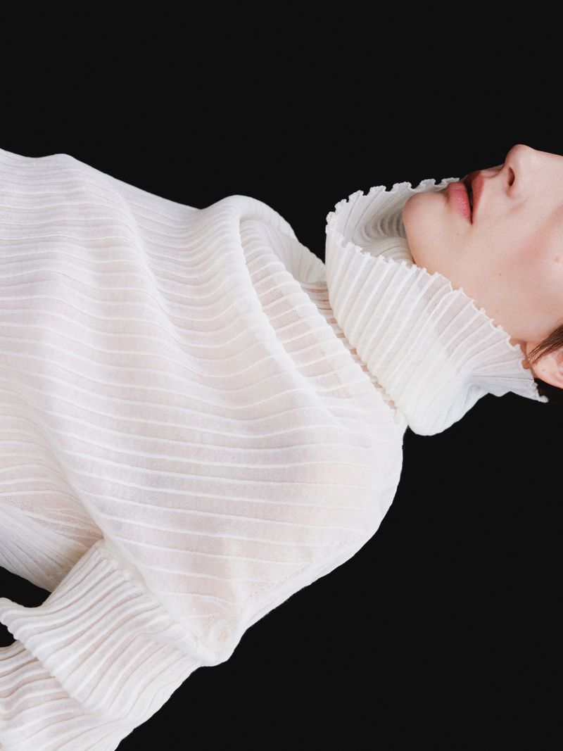 Woman in flowing white gown against black background. Close-up fashion portrait highlighting textured fabric and delicate ruffles.
