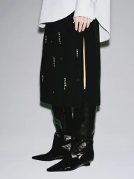 Cropped view of a model in a black embellished slit skirt, crisp white shirt, and glossy black pointed-toe boots in a studio.