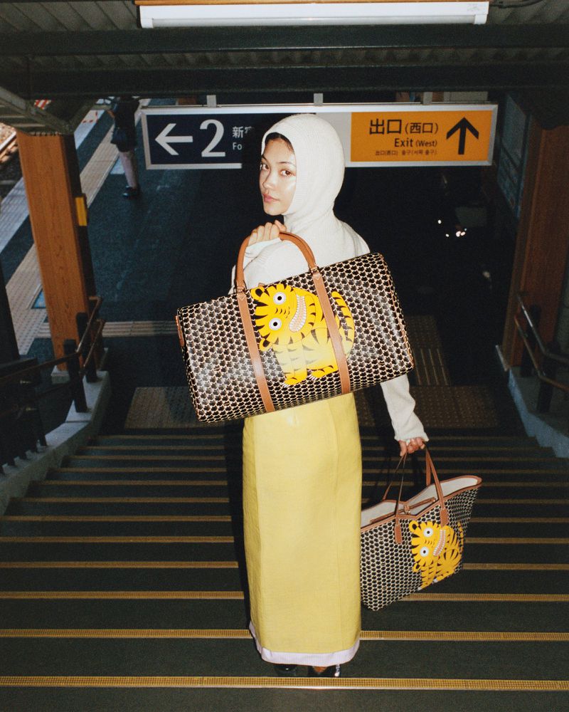 Woman in a hooded sweatshirt and yellow skirt carries a tote bag while standing on stairs in a subway station.