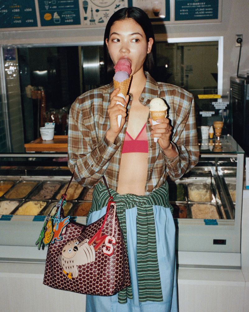 Woman in a plaid shirt enjoys ice cream cones at an ice cream parlor, holding one in each hand, with a colorful shoulder bag.