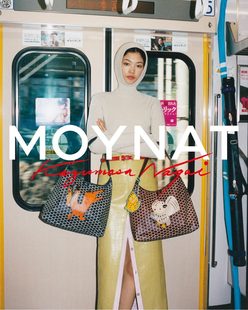 Model wearing a hooded cream top and yellow skirt standing on a train holding two patterned leather handbags