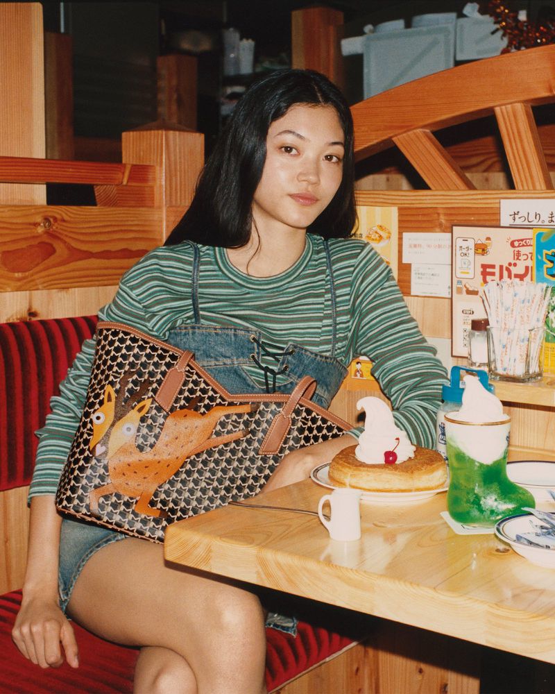Young adult woman with long dark hair poses indoors next to a table with a cake and a bag with an animal illustration.
