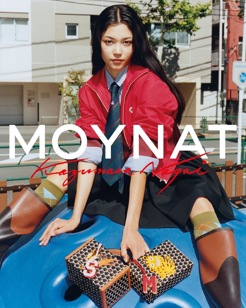 Young adult woman sits on blue play structure in a red jacket with two patterned boxes, with a city building background.