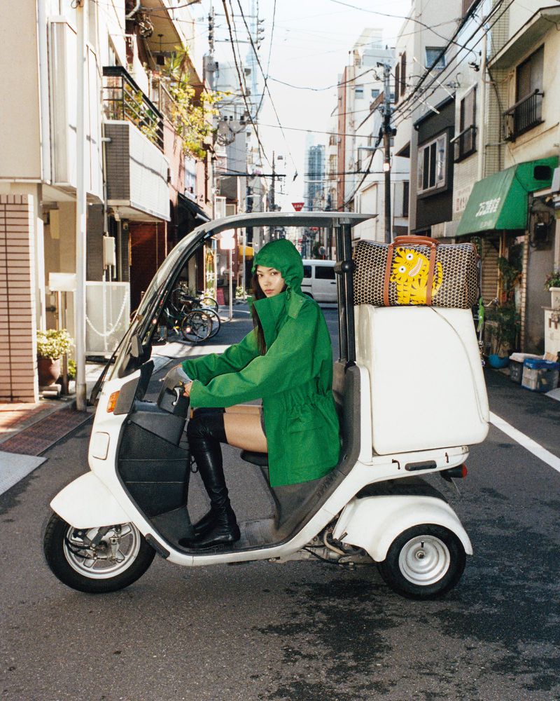 Woman in green hooded jacket rides white tricycle moped on urban street with bag. Editorial fashion shot, eye-level angle, daylight.