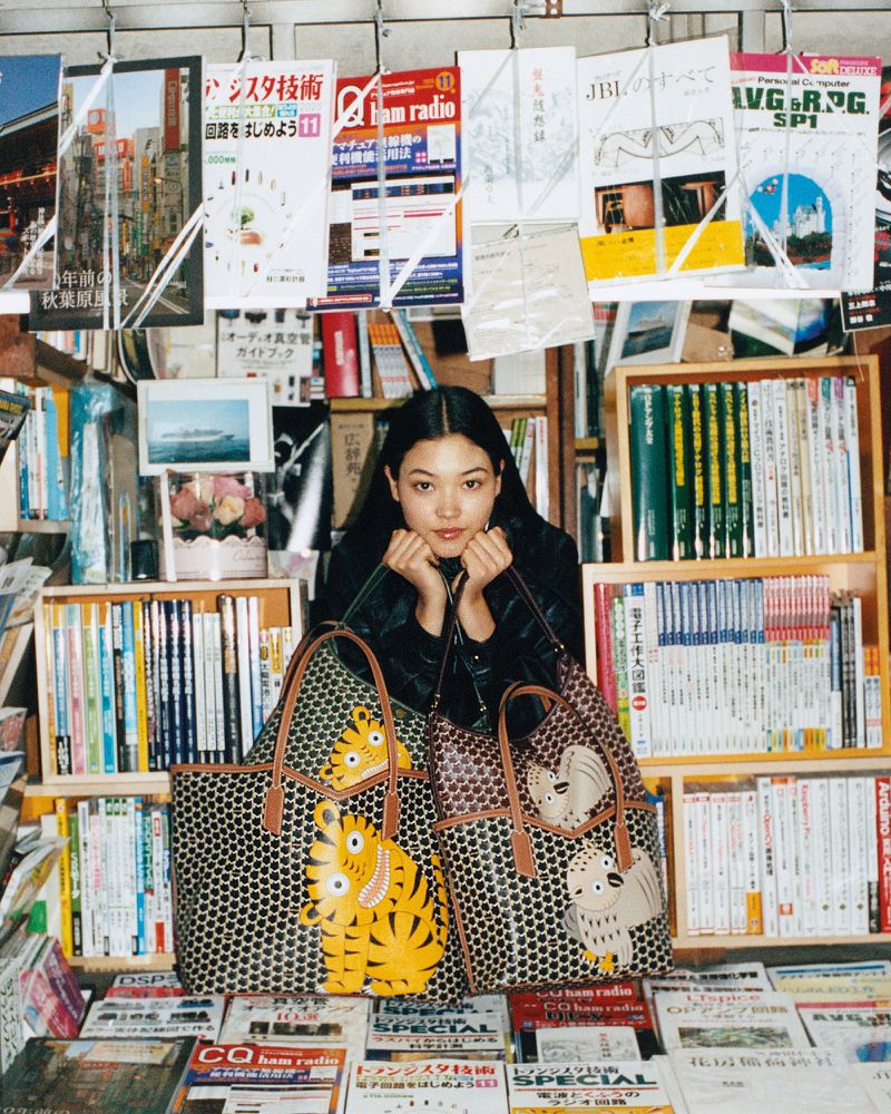 Young adult woman poses with patterned tote bags in a crowded bookstore filled with shelves and stacks of magazines and reading material.