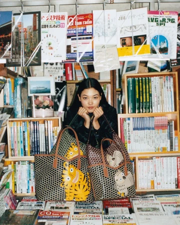 Model Mika Schneider posing behind two Moynat graphic animal tote bags surrounded by shelves and hanging magazines in a bookstore.