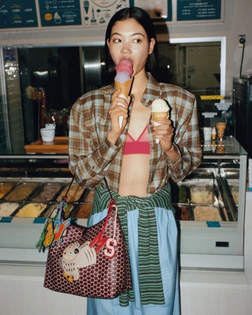 Model Mika Schneider eating an ice cream cone while carrying a patterned Moynat shoulder bag inside a gelato shop.