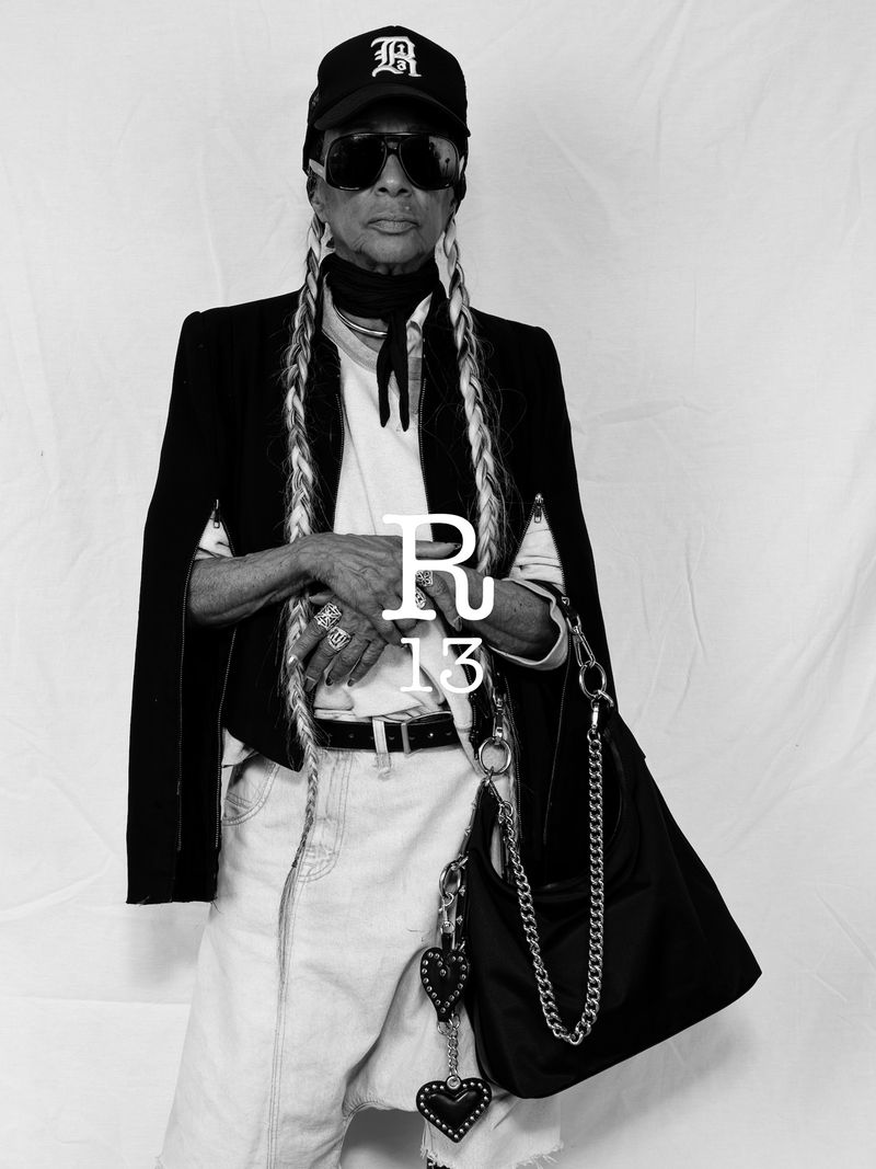 A stylish elderly woman with long braids poses in a studio wearing a baseball cap, blazer, white top, jeans, jewelry, and a black purse.