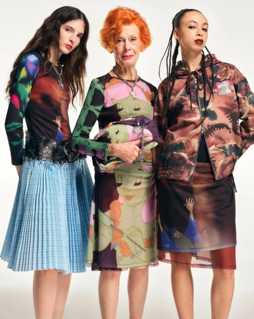 Three women of different ages stand in front of a white background, wearing colorful patterned outfits and jewelry, in a studio shot.