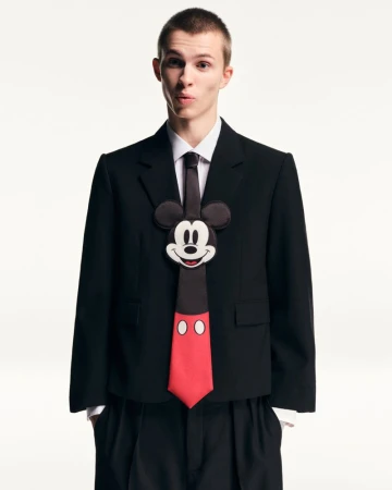 Young adult male wearing a black suit jacket with a Mickey Mouse tie against a white backdrop in a studio shot. Fashion and entertainment.