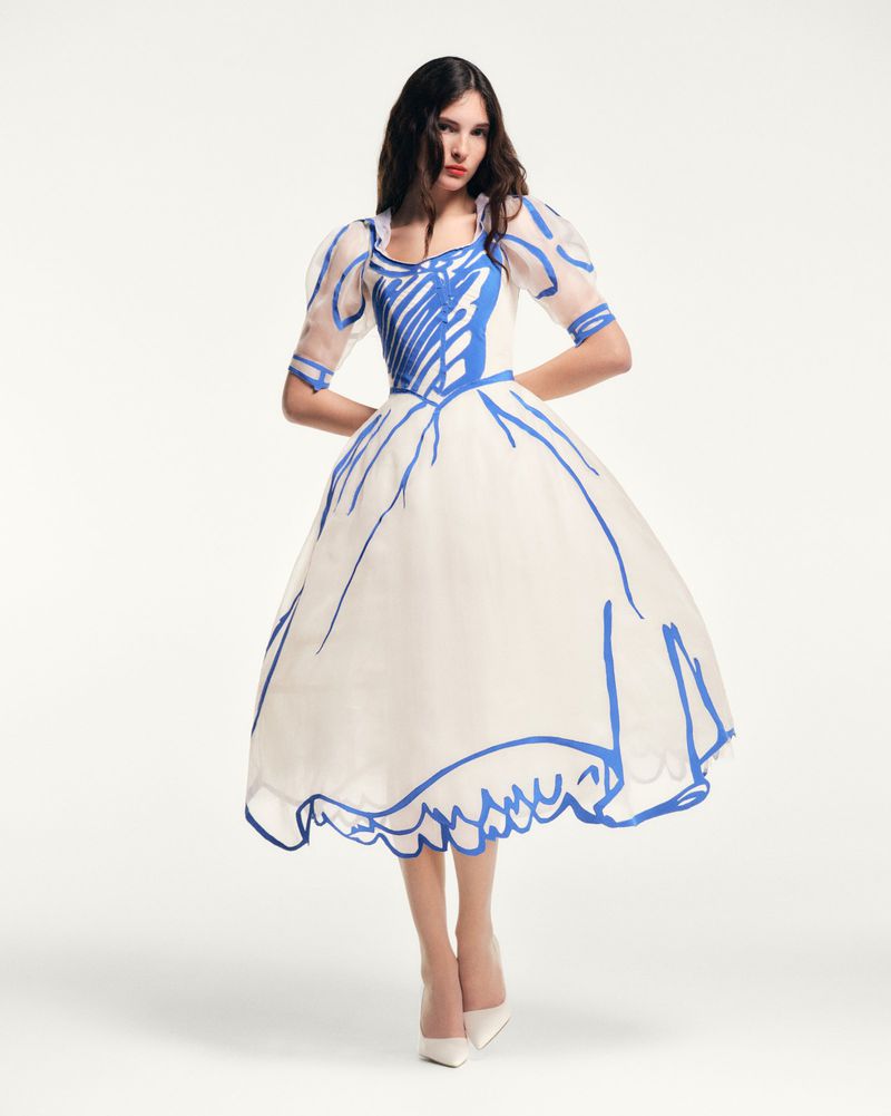 Young woman in a full, white gown with a blue drawn pattern posing in a studio with white stiletto heels for a fashion shot.