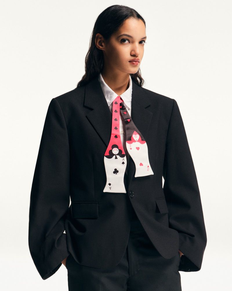A woman in a black suit and white shirt, wearing a tie with playing card designs, stands against a white backdrop in a studio setting.
