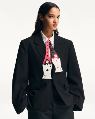 A woman in a black suit and white shirt, wearing a tie with playing card designs, stands against a white backdrop in a studio setting.
