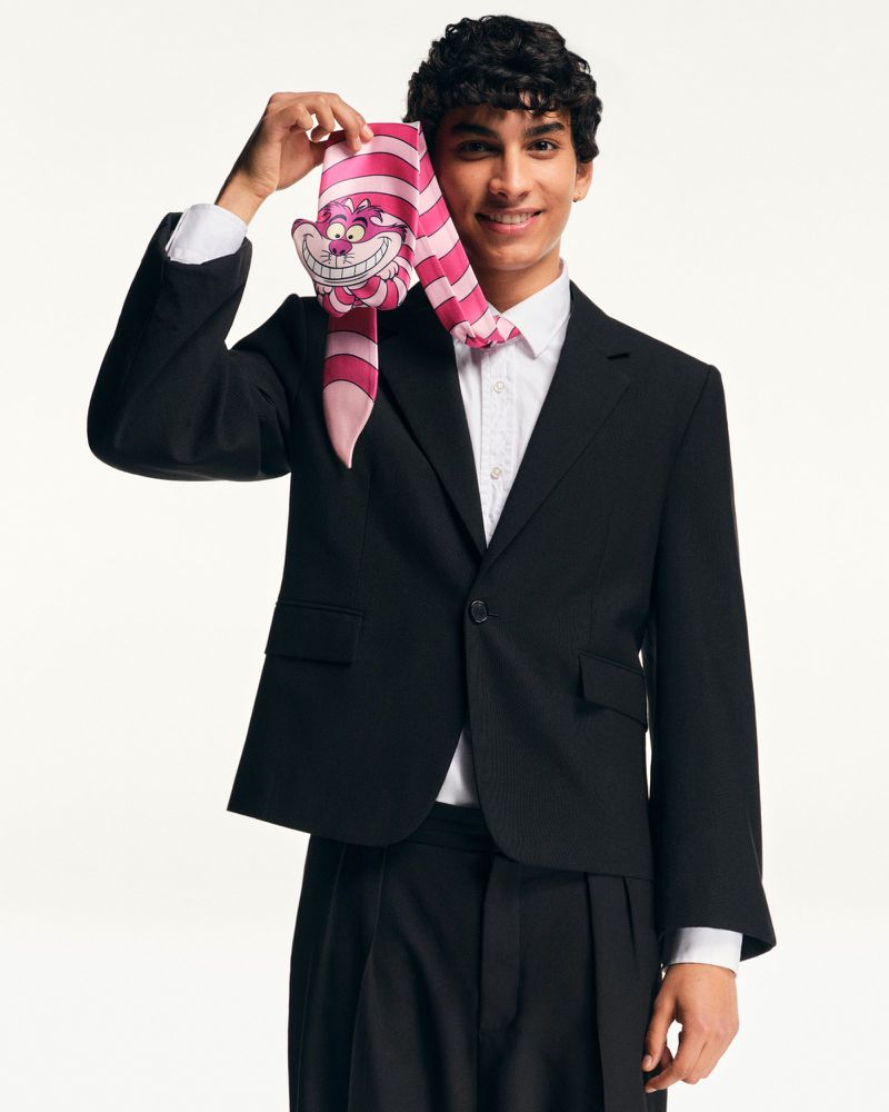 A young adult male smiles while holding a pink and white striped scarf featuring the Cheshire Cat in front of a white background.