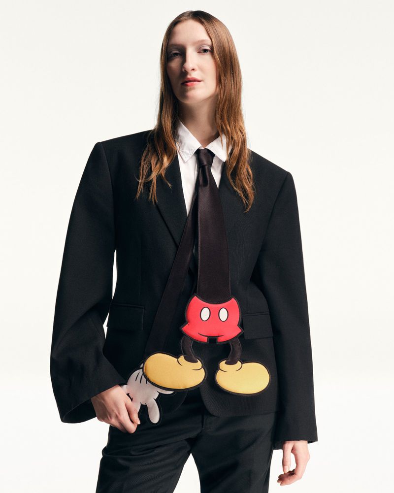 Adult woman in a black suit with a Mickey Mouse graphic, posing in a photo studio against a bright white backdrop. Fashion editorial.