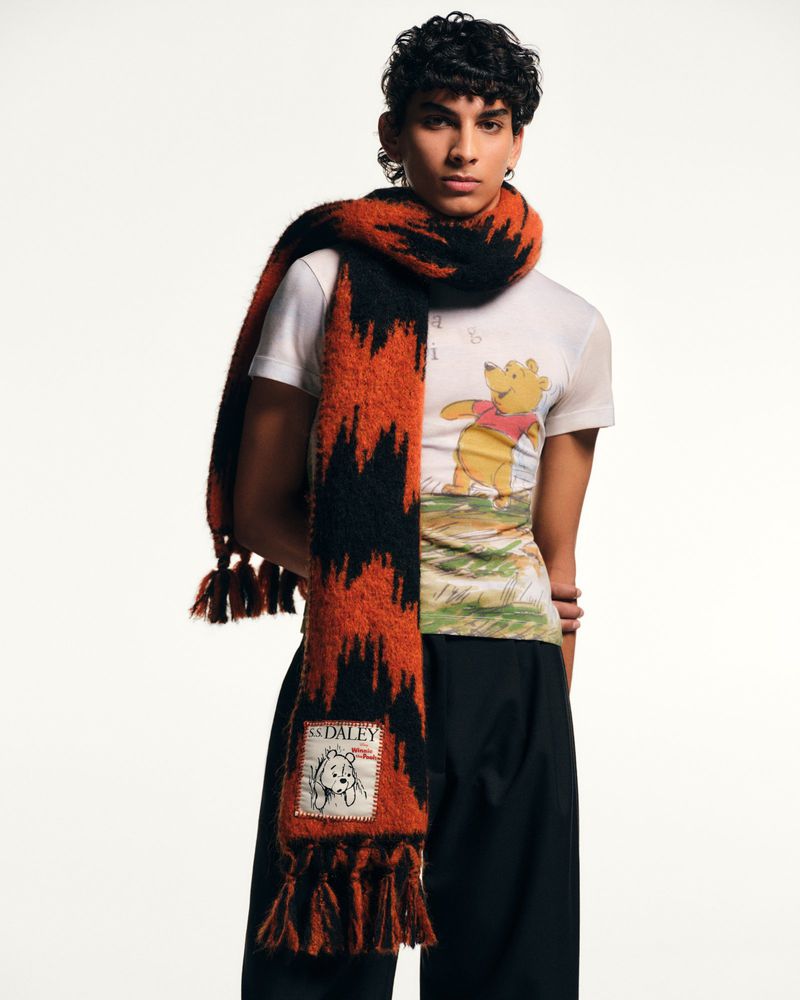 Man in white t-shirt with Winnie the Pooh graphic print and orange and black zig-zag patterned tassel scarf in a studio shot.