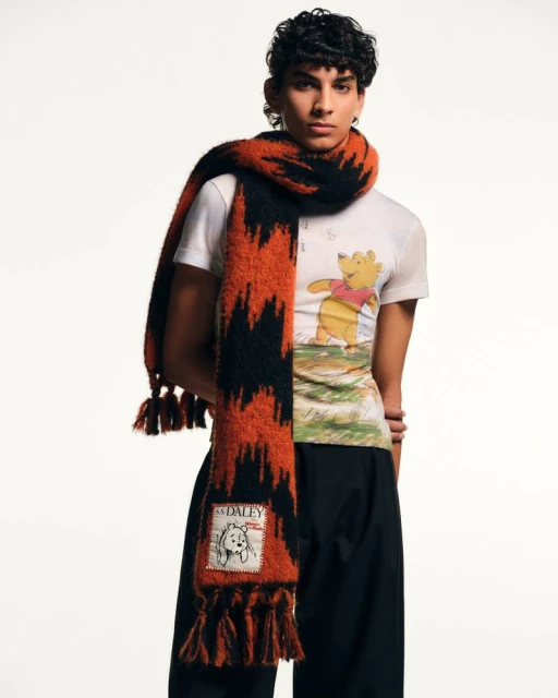 Man in white t-shirt with Winnie the Pooh graphic print and orange and black zig-zag patterned tassel scarf in a studio shot.