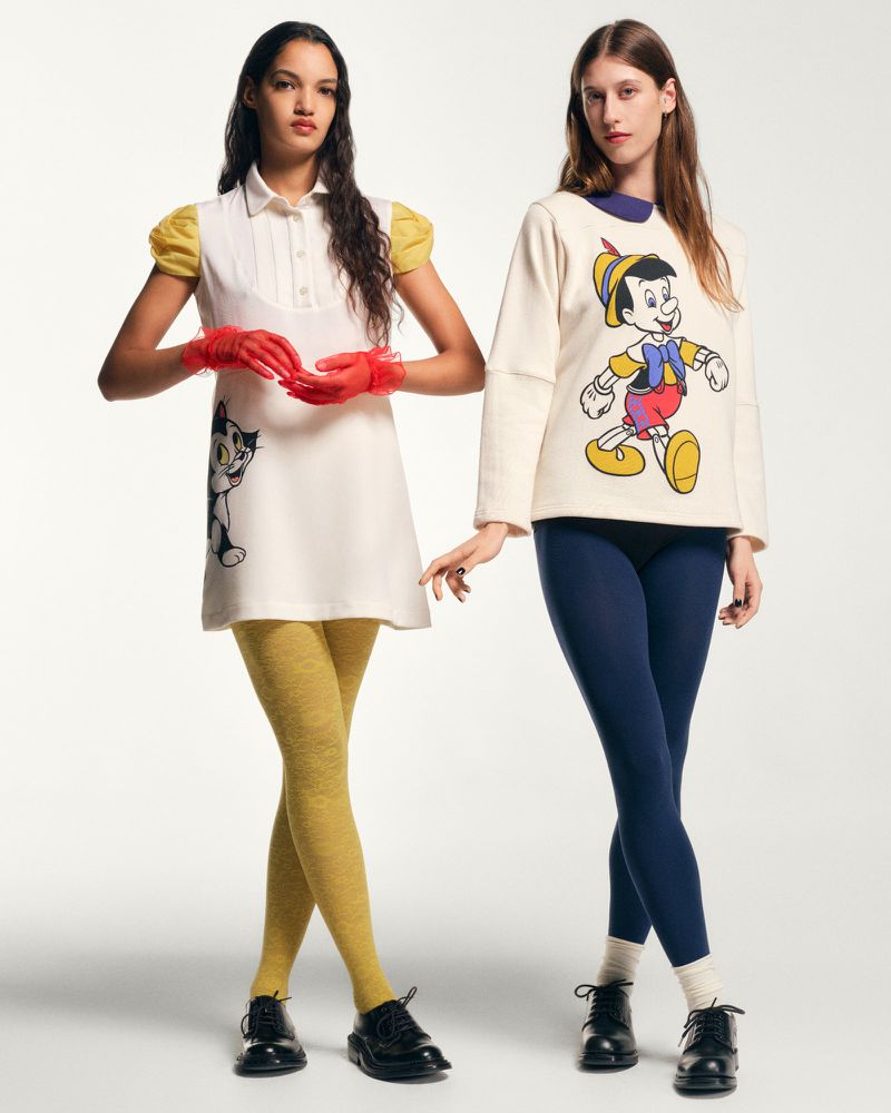 Two fashion models pose in a photo studio wearing clothing featuring cartoon characters and black patent leather shoes against a white backdrop.