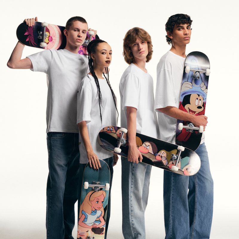 Group of four young adult models holding skateboards with Disney characters, wearing white T-shirts and jeans on a white backdrop.