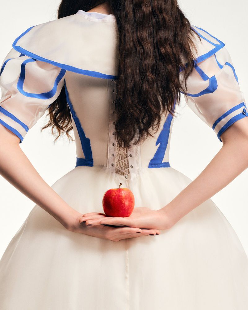 A woman wearing a white and blue dress, stands against a light background, holding a red apple behind her back. Fashion.