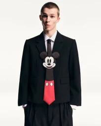 Model Enza Khoury wearing a black tailored suit jacket, white shirt, and Mickey Mouse novelty tie against a white backdrop.