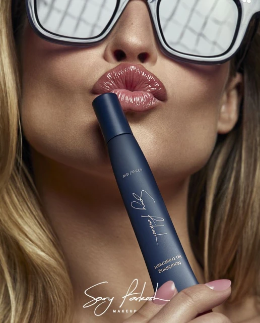 Close-up of a woman with blonde hair, wearing reflective sunglasses, holding a blue lip treatment product, with glossy lips.