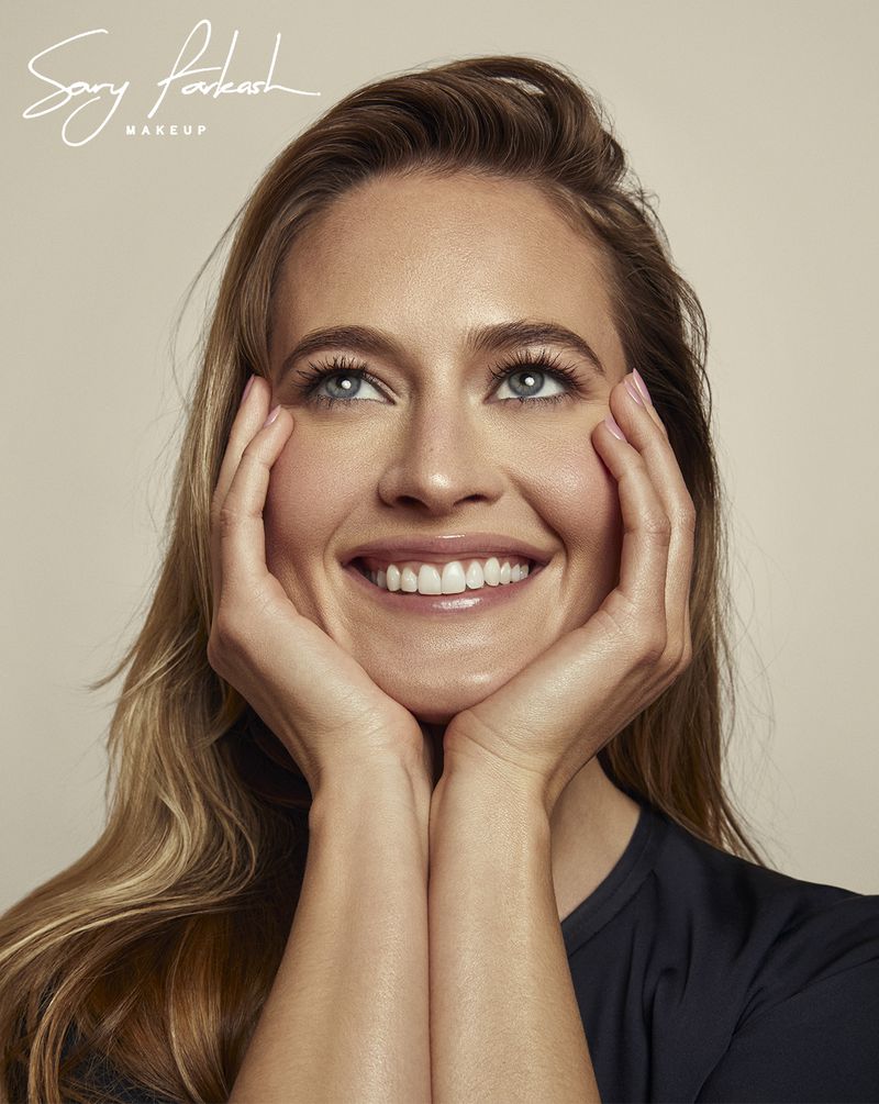 Smiling woman with hands on face looking up, wearing a black shirt, with long light brown hair and fair skin in a close-up shot.