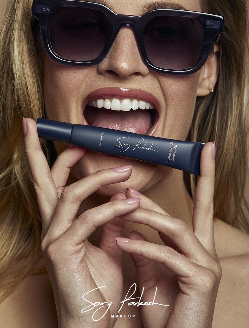 Close-up portrait of a woman wearing sunglasses holding a dark blue Sary Farkash lip treatment tube horizontally between her teeth.