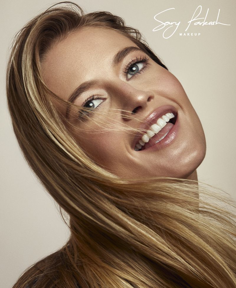 Close-up of a smiling woman with fair skin, blonde hair, and light eye makeup. She faces the camera with her tongue slightly visible.