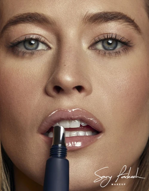 Close-up of an adult female model with fair skin, wearing lip gloss, posing while applying lip product, beauty shot, studio lighting.