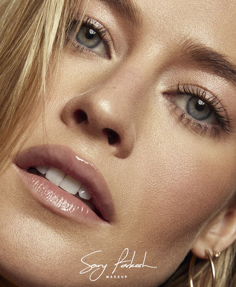 Close-up portrait of an adult female with blonde hair, blue eyes, and fair skin, emphasizing professional makeup application and gold hoop earrings.
