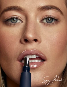 Model Hailey Carlson applying glossy lip balm with a dark blue applicator tube in a beauty close-up for Sary Farkash Makeup.