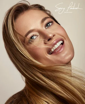 Close-up beauty portrait of model Hailey Carlson with glowing makeup and flowing blonde hair smiling against a beige studio backdrop.